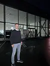 En mann står foran et glassfasadert bygg på en mørk og våt kveld.